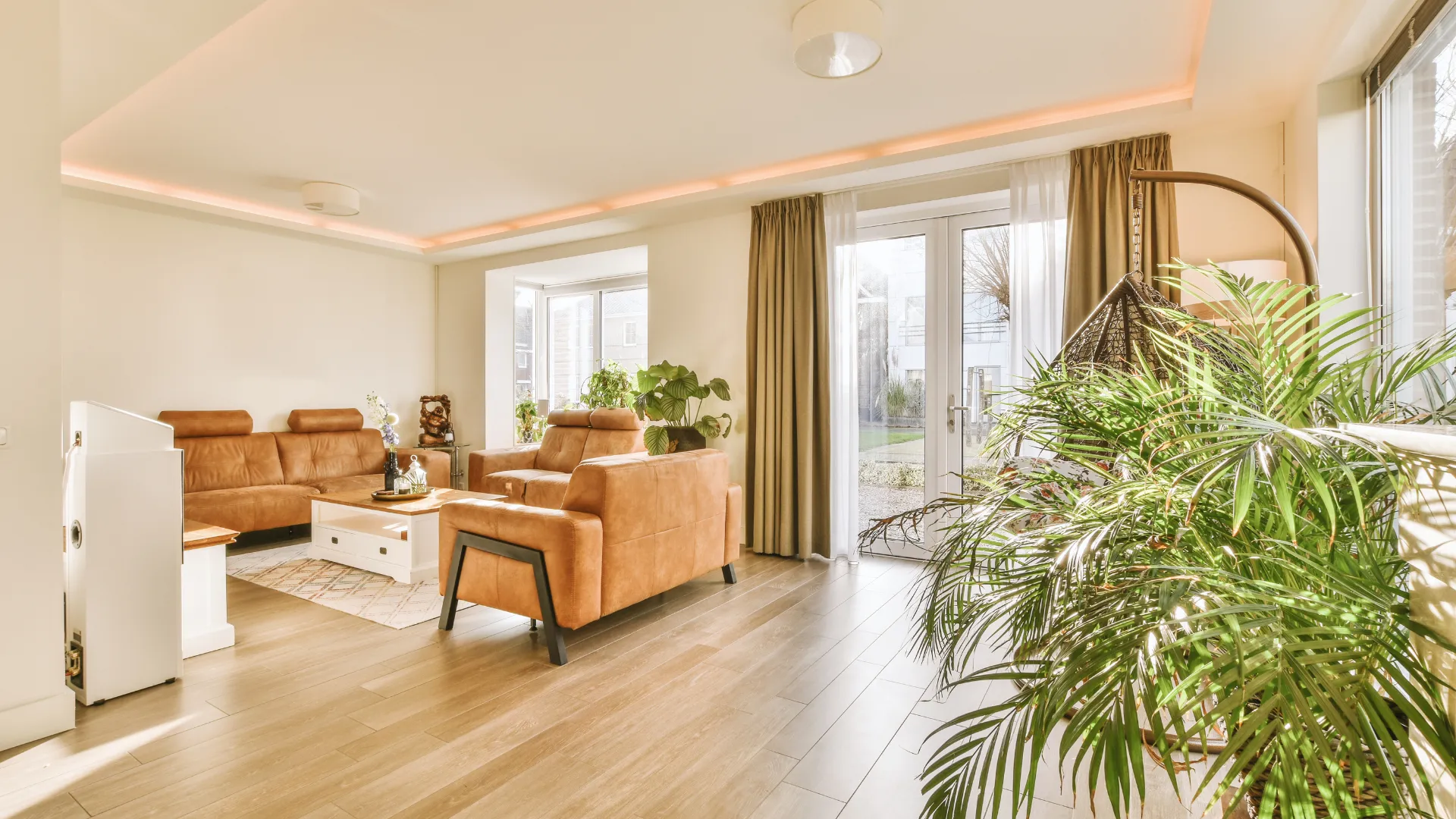 Living room with brown sofas, white table, plants, wood floor, and large windows.
