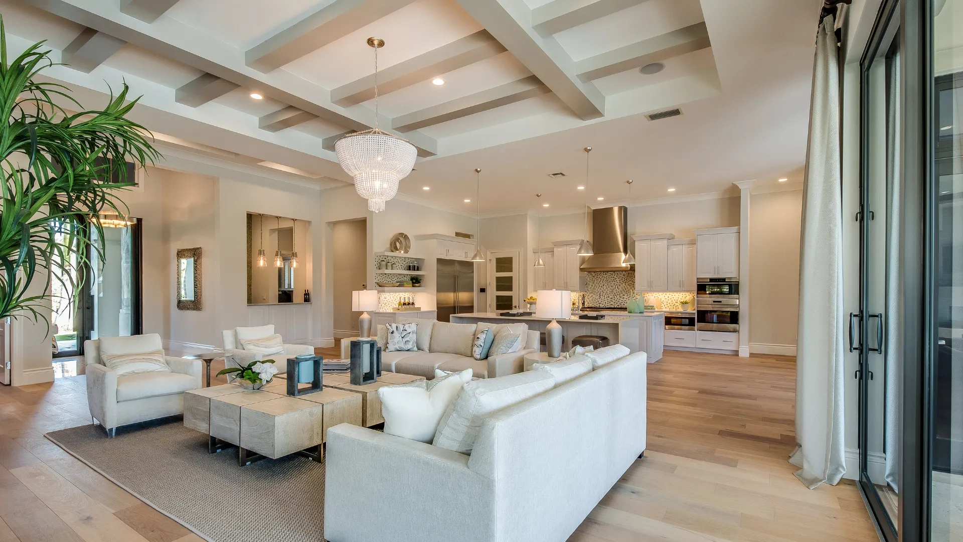 Living room with seating, large windows, plants, lights, and a kitchen in the background.