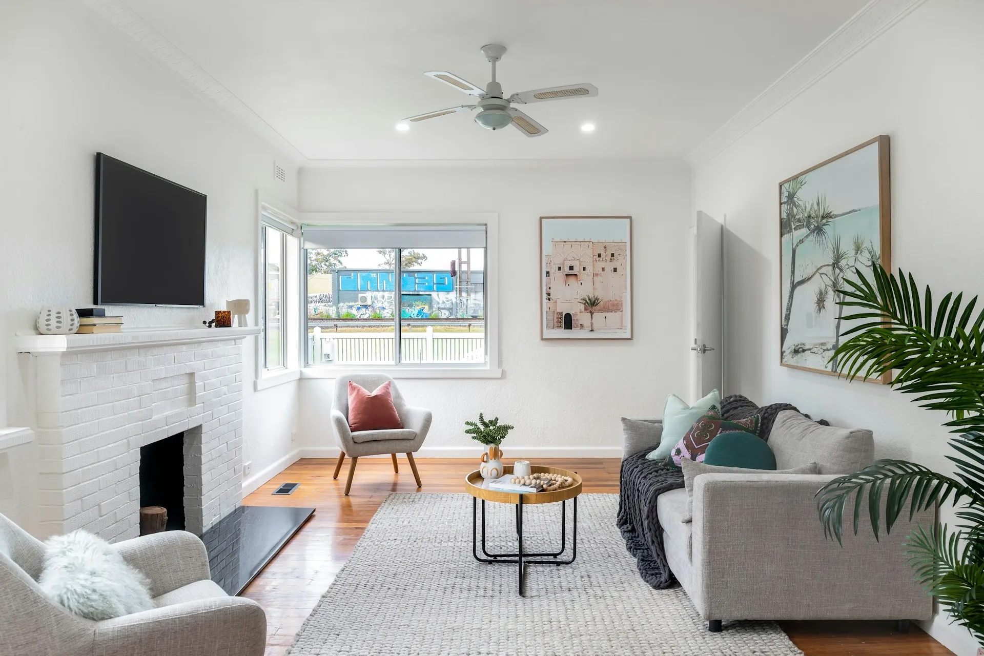 Living room with a white brick fireplace, wall TV, gray sofa, chair, coffee table, plant, and wall art.